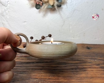 May include: A ceramic candle holder with a handle, in a light brown color, with a small white tea light candle burning inside.