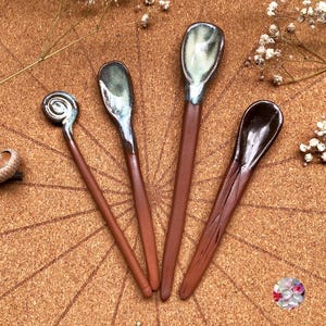 May include: Four handcrafted ceramic spoons with brown wooden handles. The spoon heads feature a variety of colors, including green, brown, and silver. The spoons are arranged on a brown surface, with a decorative acorn and flowers.