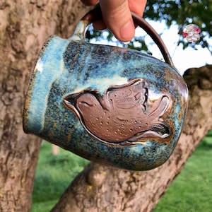 Op de afbeelding: Een blauwe en bruine keramische mok met een reliëfdesign van een vogel in de vlucht. De mok heeft een gespikkelde glazuur en een rustieke uitstraling.