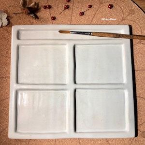 Könnte beinhalten: Weiße Keramik-Farbpalette mit vier quadratischen Vertiefungen und einer langen, dünnen rechteckigen Vertiefung zum Halten eines Pinsels. Die Palette liegt auf einer Korkoberfläche.