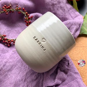Könnte beinhalten: Eine handgefertigte Keramik-Tasse mit zweifarbigem Design. Die untere Hälfte ist cremefarben beige, während die obere Hälfte hellgrau ist. Der Name "SABRINA" ist auf die Tasse geprägt, zusammen mit einem kleinen Herzsymbol. Die Tasse steht auf einem lila Stoff.