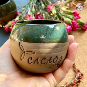 Op de afbeelding: Een handgemaakte keramische cacao-beker met een tweekleurig glazuur. De bovenste helft is glanzend teal, en de bodem is matbruin. Het woord "CACAO" is in de zijkant van de beker geëtst, met een bloemmotief. De beker wordt in een hand gehouden.