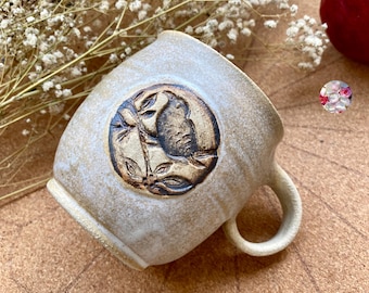 May include: A handmade ceramic mug with a beige glaze and a circular, brown embossed design featuring a bird and leaves. The mug has a rounded handle and a textured surface, perfect for enjoying coffee or tea.
