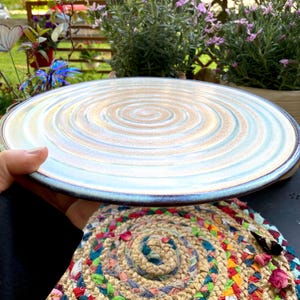 May include: A large, round ceramic plate with a spiral design in shades of blue, tan, and white. The plate has a dark blue rim and is held by a person. The plate is likely used for serving food.