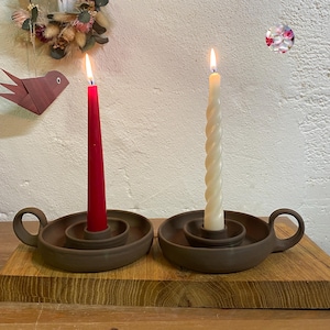 May include: Two brown ceramic candle holders with handles, each holding a lit candle. One candle is red, the other is a twisted ivory color. The holders sit on a wooden surface.