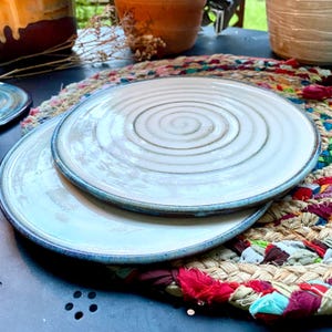 Op de afbeelding: Twee witte keramische borden met een spiraalvormig ontwerp en blauwe randen zijn gestapeld op een kleurrijke geweven placemat. De borden hebben een glanzende afwerking en staan op een donkere ondergrond, wat duidt op een eetgelegenheid.