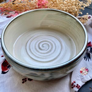 May include: A handmade ceramic bowl with a cream-colored interior and a spiral design in the center. The bowl has a dark green rim and a light green exterior. The bowl is sitting on a floral patterned cloth.