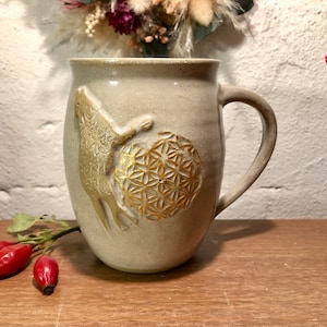 May include: A ceramic mug with a beige glaze and a gold-colored design of a stylized animal and a geometric pattern.