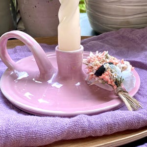 Può includere: Un portacandele in ceramica rosa chiaro lucido con un manico curvo e un supporto centrale che ospita una candela affusolata ritorta color crema. Un piccolo mazzetto decorativo di fiori secchi rosa e bianchi è fissato alla base del supporto.
