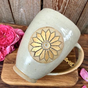 May include: A ceramic mug with a white glaze and a gold-coloured floral design. The mug has a handle and is sitting on a wooden surface.