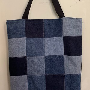 May include: A patchwork tote bag made from different shades of blue denim. The bag has black straps and a rectangular shape.