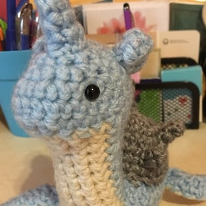 Puede incluir: Un animal de peluche azul y blanco tejido a crochet con una concha gris. El animal tiene un hocico largo y puntiagudo y un ojo grande y redondo.