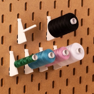May include: A pegboard with white hooks holding spools of thread in various colours, including black, green, blue, pink, and white.
