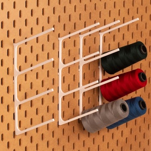 以下が含まれることがあります： 白いプラスチック製の壁掛け収納ラックに、緑、赤、グレー、青の4色の糸巻きが置かれています