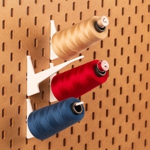 May include: Three spools of thread are hanging on a white pegboard. The spools are red, blue, and beige. The pegboard has a grid pattern.