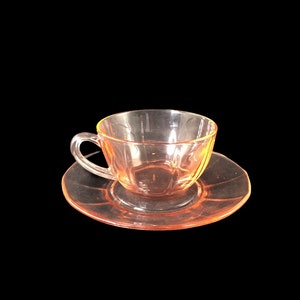 May include: A pink glass teacup and saucer set. The cup and saucer are both clear glass with a light pink tint.