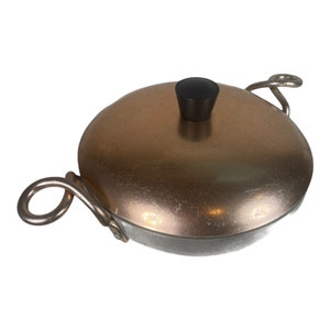 May include: A vintage copper-toned aluminum pot with two handles and a black knob on the lid.