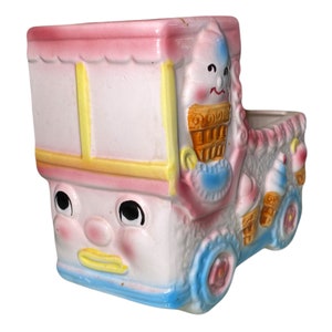 May include: A vintage ceramic ice cream truck planter with a pink, white, and blue color scheme. The truck has a smiling face and a cone of ice cream on top.