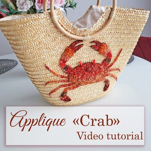 May include: A straw tote bag with wooden handles and a red beaded crab applique. The bag is on a white surface with a floral arrangement in the background. The text "Applique «Crab» Video tutorial" is at the bottom of the image.