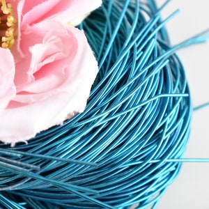 May include: A close-up of a spool of turquoise blue wire. The wire is tightly wound and has a shiny finish. A pink flower is partially visible in the background.