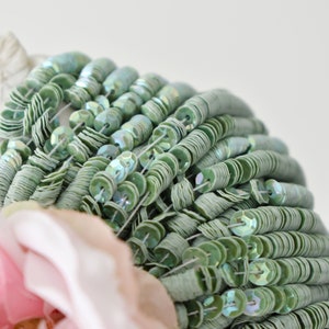 Pode incluir: Um close-up de uma linha de lantejoulas verde claro iridescentes. As lantejoulas são costuradas juntas em uma linha contínua e são organizadas em um padrão circular.