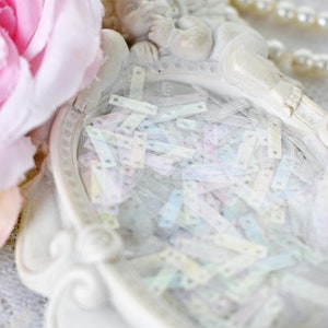 May include: A white porcelain cherub figurine with wings, lying on its side, with a white ornate frame around a pile of iridescent sequins.