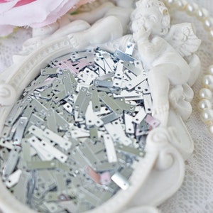 Puede incluir: Una figurita de ángel de cerámica blanca con alas, sosteniendo un pequeño corazón de cerámica blanca. La figurita está sentada en una bandeja de cerámica blanca llena de piezas de metal plateado.