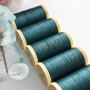 May include: A row of six spools of teal green thread with cream colored labels. The thread is wound tightly on the spools.