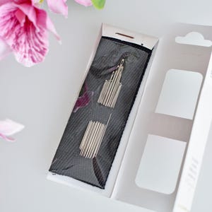 May include: A close-up of a package of needles for sewing. The needles are silver and are packaged in a black and white plastic case. The case is open and the needles are visible.