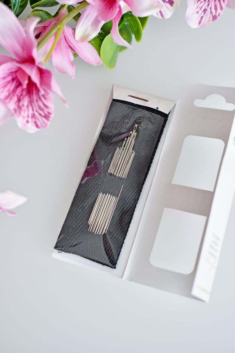 May include: A close-up of a package of needles for sewing. The needles are silver and are packaged in a black and white plastic case. The case is open and the needles are visible.