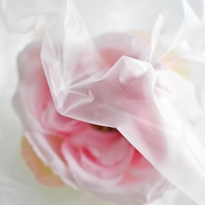 Puede incluir: Una delicada flor rosa está envuelta en una bolsa de plástico transparente blanca. La flor es parcialmente visible a través del plástico, creando un efecto suave y etéreo.
