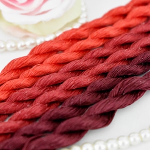 Può includere: Cinque matasse di filo da ricamo in un gradiente di colori rosso, arancione e bordeaux. Il filo è disposto a treccia.