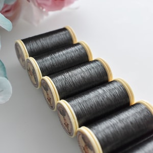 Puede incluir: Seis carretes de hilo negro con etiquetas de color crema. El hilo está enrollado alrededor de los carretes y las etiquetas tienen un diseño vintage.