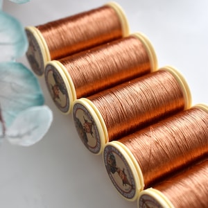 May include: A close-up of six spools of copper-coloured thread. The spools are arranged in a row, with the labels facing the viewer. The labels feature a vintage-style illustration of a woman with a sewing machine.