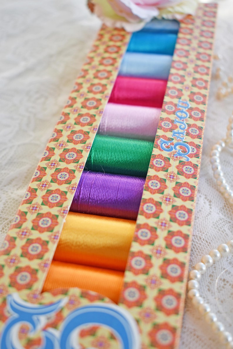 May include: A box containing 10 spools of colourful thread. The spools are arranged in a row, with the colours ranging from blue to orange. The box is decorated with a floral pattern and the text "50 Colours".