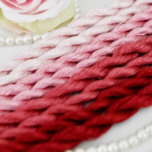 May include: A collection of six embroidery floss strands in a gradient of pink and red shades. The floss is braided and arranged in a fan shape. The floss is on a white surface with a pearl necklace.