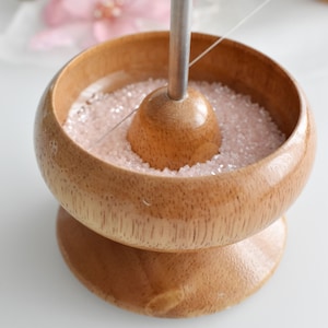 May include: A wooden beading bowl with a metal needle and pink beads. The bowl has a base and a round top with a hole in the centre for the needle.