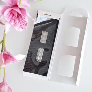 May include: A white box with a black fabric insert containing a set of silver sewing needles. The box is open and the fabric insert is visible.