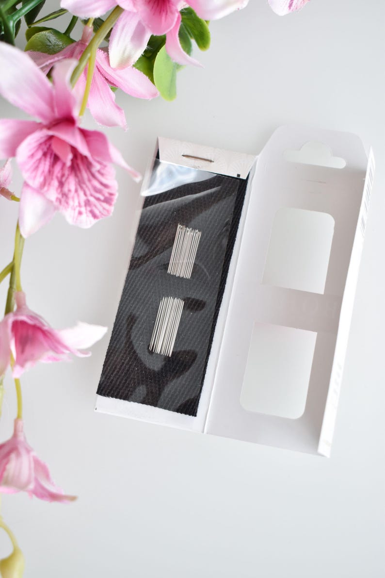 May include: A white box with a black fabric insert containing a set of silver sewing needles. The box is open and the fabric insert is visible.