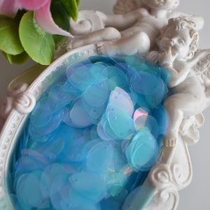 Può includere: Un contenitore decorativo bianco a tema cherubino pieno di paillettes blu iridescenti. Il contenitore ha una forma ovale con dettagli ornati. Fiori rosa e viola sono visibili sullo sfondo.