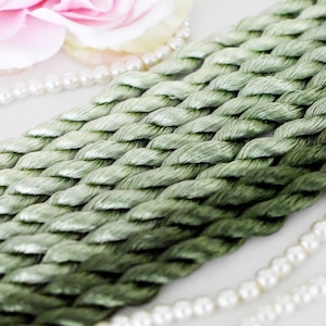 Puede incluir: Primer plano de un cordón trenzado verde claro, posiblemente hilo de bordar o cuerda decorativa. Una flor rosa y un collar de perlas blancas están en el fondo. El cordón tiene una textura tejida y diferentes tonos de verde.