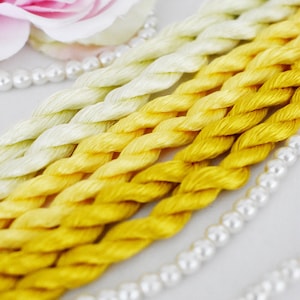 Peut inclure: Quatre écheveaux de fil à broder dans des tons de jaune et de vert. Le fil est disposé en dégradé du vert clair au jaune foncé. Le fil est posé sur une surface blanche avec une chaîne de perles blanches.