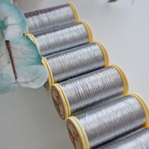 Op de afbeelding: Een close-up van zes spoelen zilveren metallic garen. De spoelen zijn in een rij gerangschikt, met de uiteinden van het garen naar de kijker gericht. De spoelen zijn allemaal even groot en hebben een gele kunststof basis.