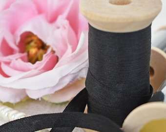 Pode incluir: Uma bobina de madeira com fita preta está colocada perto de uma rosa rosa e um colar de pérolas. A fita está enrolada na bobina. Outras bobinas de madeira e fitas estão no fundo.