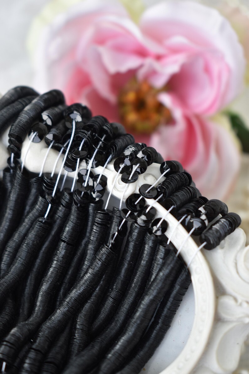 May include: Black sequins and black metallic beads strung on white thread. The beads are arranged in a circle, with the sequins in the center.