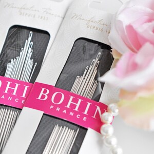 May include: Two packages of Bohin sewing needles, a French brand, with the text "Manufacture Francaise Depuis 1822" on the packaging. The needles are silver and are arranged in a black background. The packages are on a white surface with a pearl necklace and a pink flower.