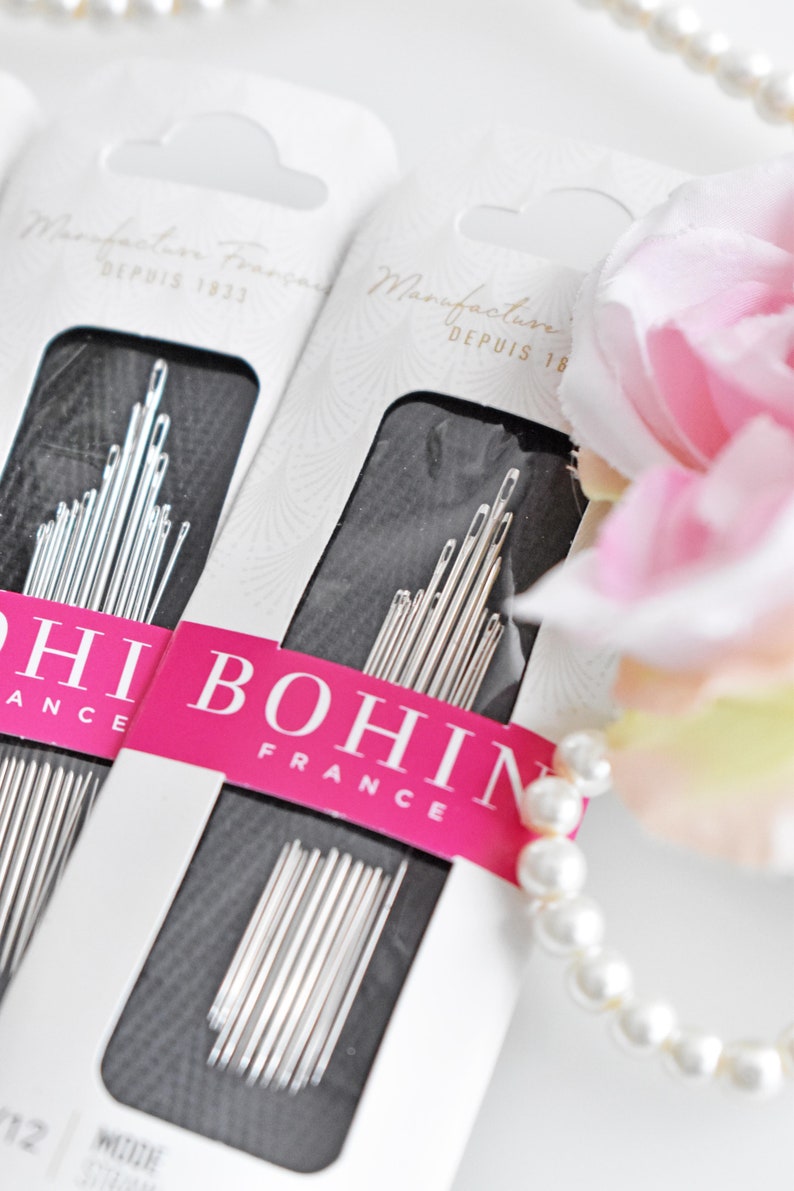 May include: Two packages of Bohin sewing needles, a French brand, with the text "Manufacture Francaise Depuis 1822" on the packaging. The needles are silver and are arranged in a black background. The packages are on a white surface with a pearl necklace and a pink flower.