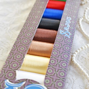 May include: A box containing eight spools of thread in a variety of colours, including white, gold, brown, black, blue, red, green, and white. The box has a purple and green patterned design with the text "El Gato" on the side.