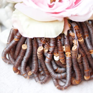 May include: Close-up of a bundle of brown sequins, strung together, with a light pink and white flower in the background. The sequins are circular and have a metallic sheen. The overall aesthetic is delicate and decorative.