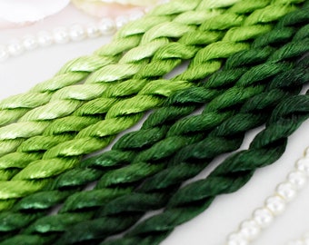 May include: A collection of six strands of embroidery floss in shades of green, ranging from light green to dark green. The floss is arranged in a gradient pattern, with the lightest shade at the top and the darkest shade at the bottom.